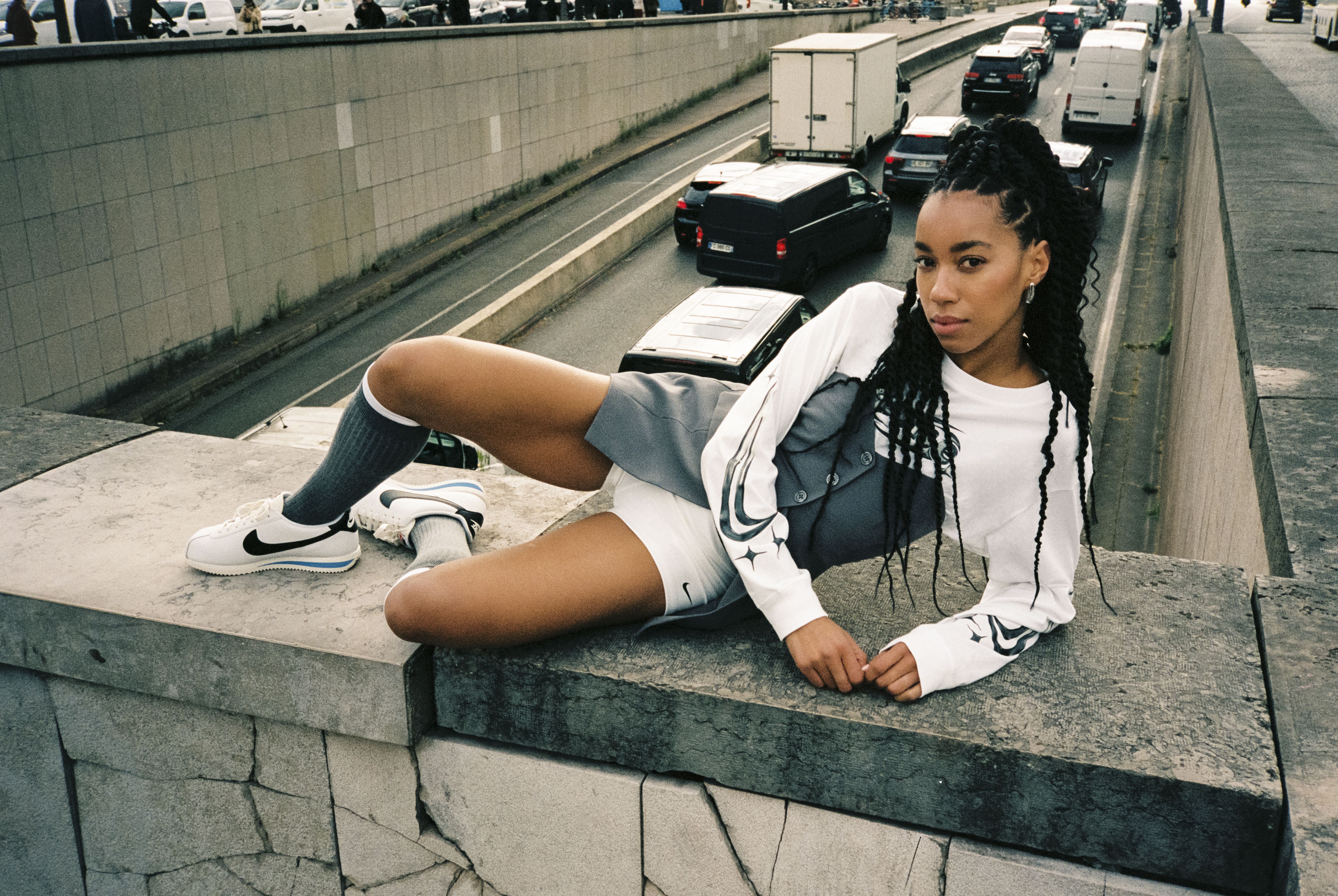 Жена в бял суичър на NIKE и бели NIKE обувки, позираща на стена на оживена улица